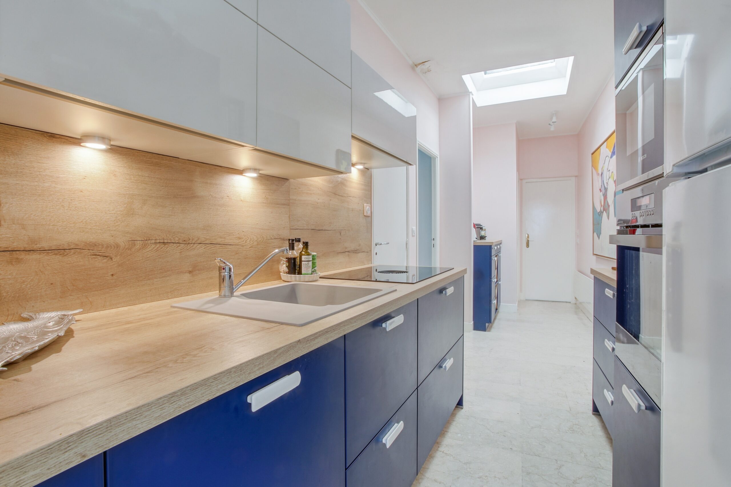 Cuisine longue avec armoires bleues, comptoir bois et plaques induction, adaptée à un appartement