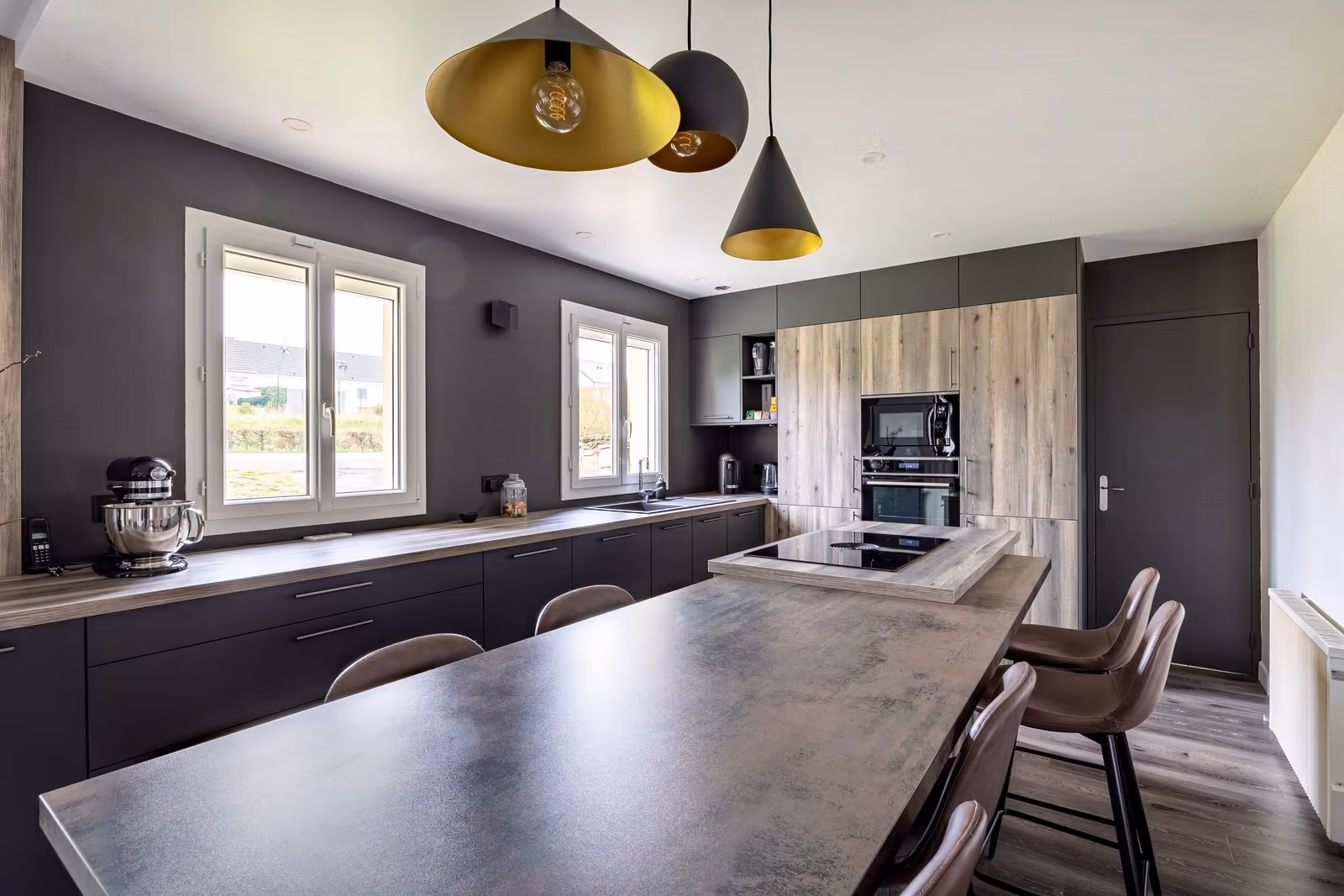 Grande cuisine ouverte avec armoires anthracite, plan de travail béton, colonne en bois et suspension dorée